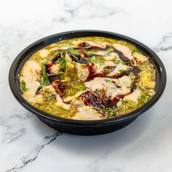 Indian chaat with green and white sauces, dark chutney, and crispy noodles in a black bowl.