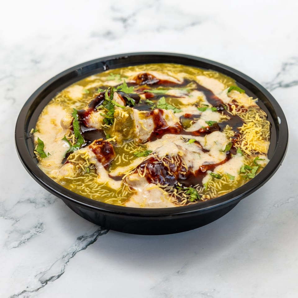 Indian chaat with green and white sauces, dark chutney, and crispy noodles in a black bowl.